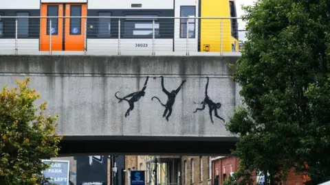 PA Media Three black monkeys appearing to hang from a grey concrete railway bridge in Brick Lane