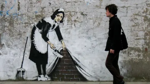 Getty Images Sweep It Under The Carpet on a wall in Camden, depicting a maid in black and white with a dustpan and brush appearing to pull up a painted wall to expose the bricks behind